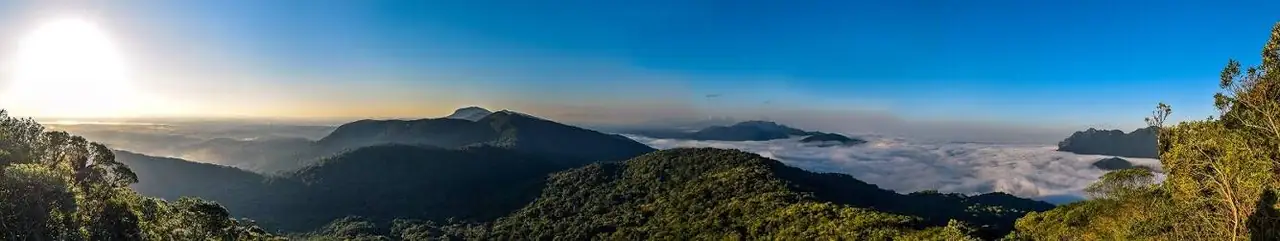 Panorama com o sol baixo à esquerda e um mar de nuvens entre as montanhas